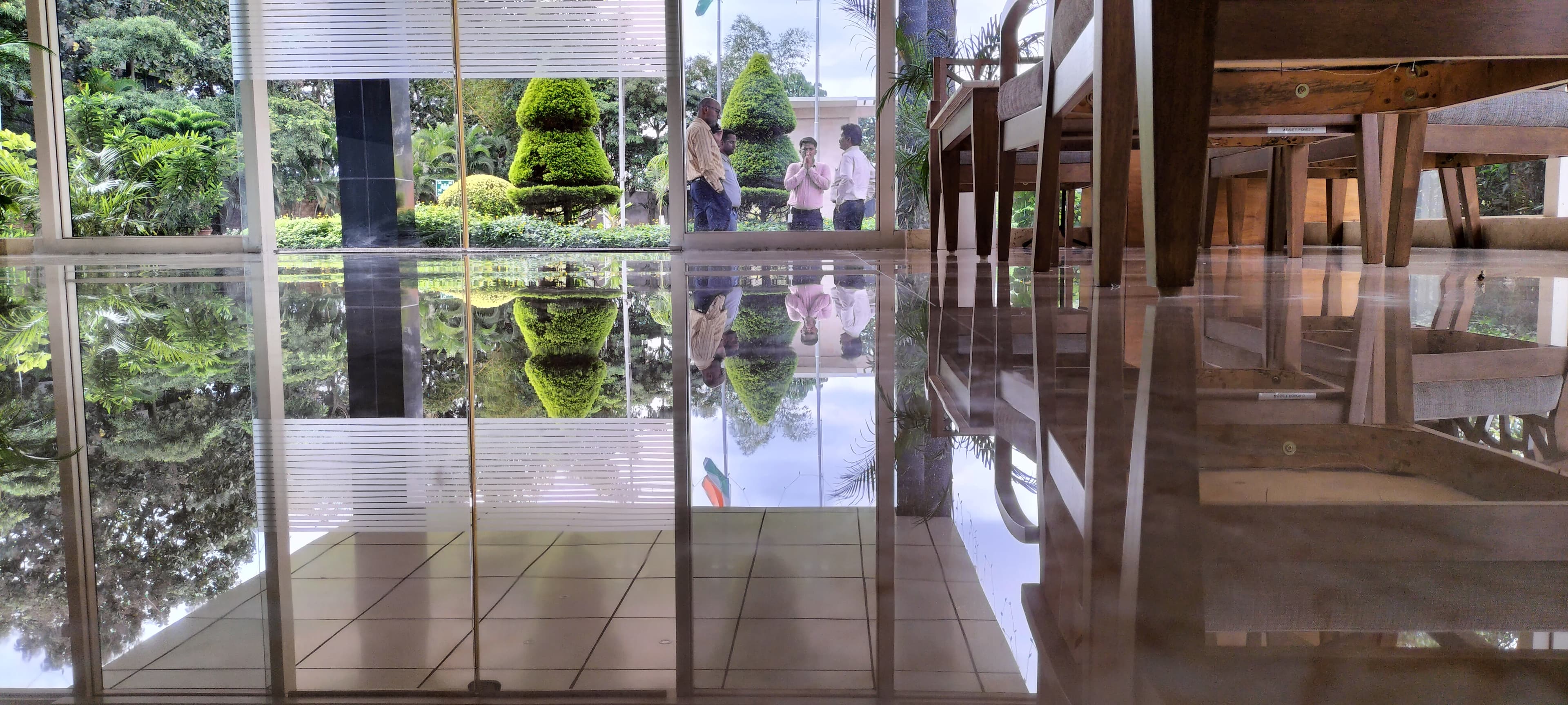 Polished floor reflecting a garden with topiary trees and men standing outside glass windows.